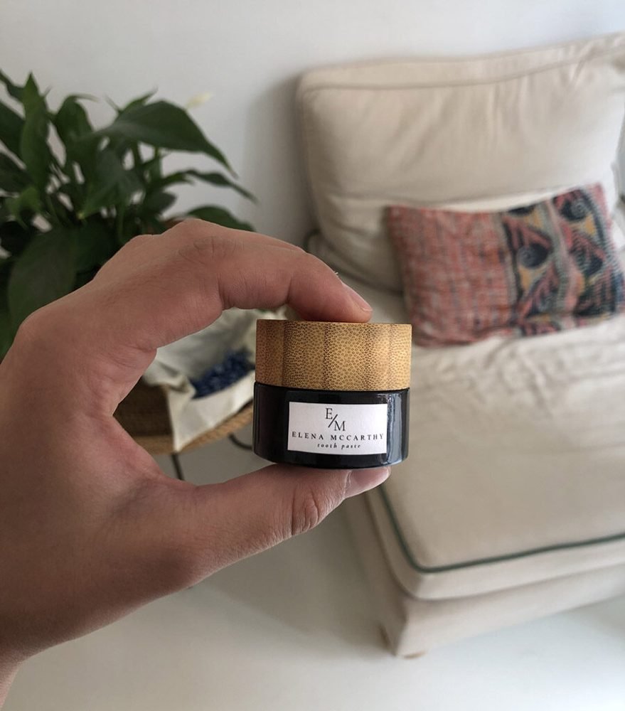 A jar of Elena McCarthy natural coconut toothpaste held in a bright living room